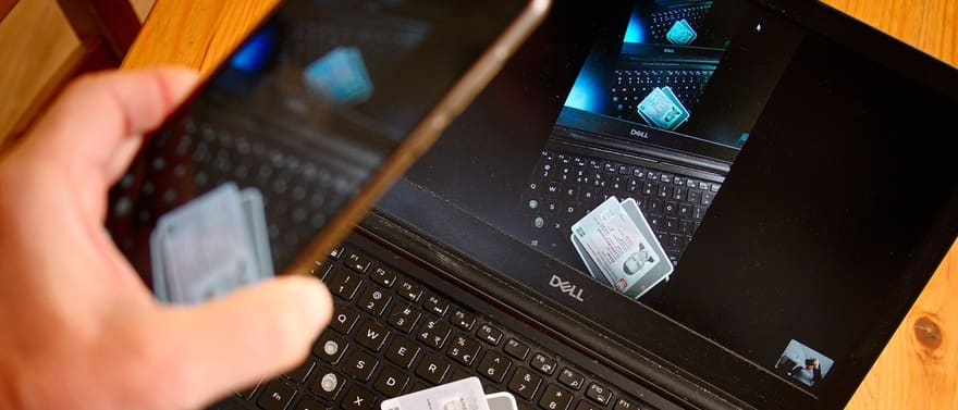 Mão segurando um celular diante de um notebook aberto, cuja tela mostra a foto de um documento de identidade.