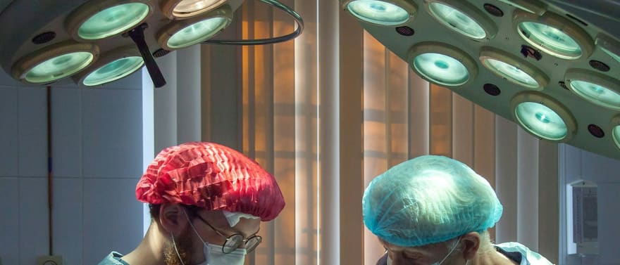 Dois cirurgiões de touca e máscara operando sob as luzes de uma sala de cirurgia.