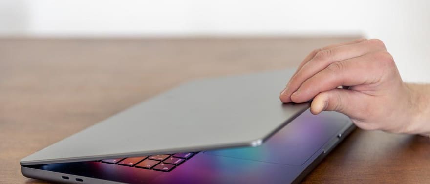 Mão fechando a tampa de um notebook prateado sobre uma mesa de madeira.