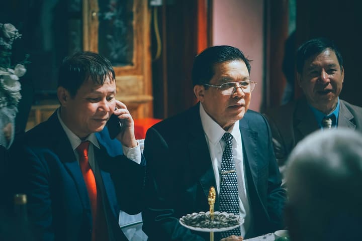 Três homens de terno sentados à mesa em um encontro formal, um deles falando ao celular.