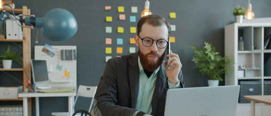 Homem de óculos falando ao celular enquanto usa um notebook, em um escritório com post-its na parede.