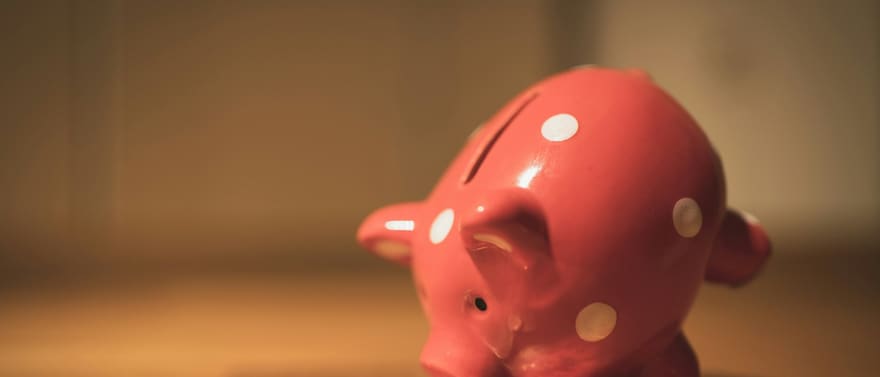 Cofrinho vermelho de bolinhas brancas com moedas espalhadas sobre uma mesa de madeira.