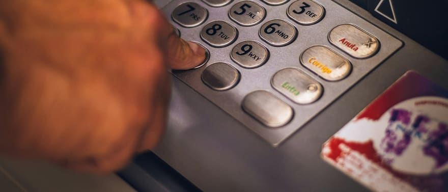 Dedo digitando a senha no teclado de um caixa eletrônico, com um cartão ao lado.